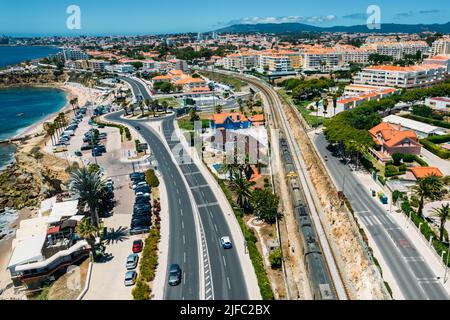 Luftdrohnenansicht des Zuges, der in der Nähe der Marginal Avenue und der Küste mit dem Parade-Viertel im Großraum Lissabon, Portugal, vorbeifährt Stockfoto
