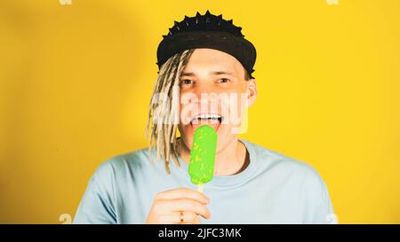 Junger, hübscher Mann mit Dreadlocks in schwarzer Mütze, der Eis auf gelbem Hintergrund leckt. Glücklicher Kerl, der Eisbecher isst, die Kamera anschaut und lächelt Stockfoto