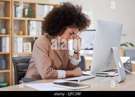 Junge afroamerikanische Geschäftsfrau, die gestresst aussieht, während sie einen Computer an ihrem Schreibtisch im Büro benutzt. Schwarze Frau mit Kopfschmerzen. Frau mit gemischter Rasse Stockfoto
