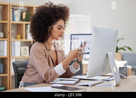Eine junge, vielbeschäftigte afroamerikanische Frau, die bei ihrem Bürojob einen Schreibtisch und ein Mobiltelefon benutzt. Schwarze Frau mit einem Afro, der sie in den sozialen Medien ancheckt Stockfoto