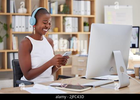Lächelnde afroamerikanische Geschäftsfrau, die per Smartphone eine Nachricht sendet oder Musik spielt, während sie kabellose Kopfhörer trägt und an ihrem pc sitzt Stockfoto