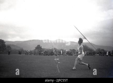 1954, historisch, mit den schottischen Bergen als Hintergrund, draußen auf einem Feld, läuft ein männlicher Junior-Athlet in Shorts und Weste, um den Speer zu werfen, Schottland, Großbritannien. Der Anlauf ist im Speer wichtig, um Schwung für den Wurf zu schaffen, was nur dann gilt, wenn die Spitze des Speers nach dem Wurf zuerst auf den Boden trifft. Stockfoto