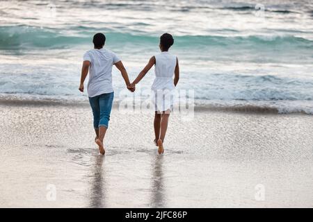 Glückliches afroamerikanisches Paar, das einen Tag am Meer zusammen verbringt. Rückansicht eines fröhlichen Freundes und einer Freundin, die am Strand ins Wasser laufen Stockfoto