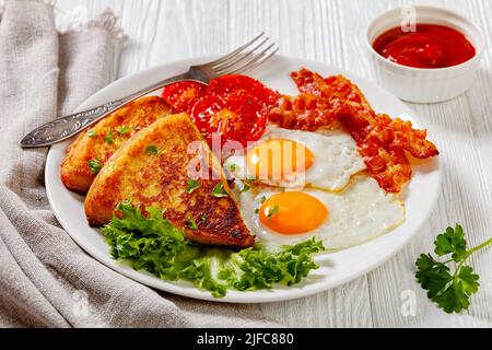 Spiegeleier, Raser- oder Speckscheiben, Kartoffelfarne, gegrillte Tomaten und frischer Salat auf weißem Teller mit Gabel auf weißem Holztisch, irland-Frühstück Stockfoto