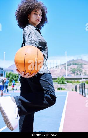 Porträt einer trendigen Mixed Race Frau mit einem Afro, der einen Basketball hält und draußen auf einem Platz posiert. Modische hispanic mit lockigen Haaren Gefühl Stockfoto