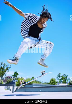 Cooler Junge, der auf seinem Skateboard im Skatepark Tricks vorführt. Fokussierter junger afroamerikanischer Mann, der springt, um sein Skateboard in die Luft zu kippen Stockfoto