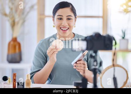 Glückliche junge Beauty-Bloggerin hält ein Beauty-Produkt und einen Schwamm hoch, während sie ein Make-up-Tutorial macht und mit ihrem Stativ Aufnahmen macht. Einflussnehmer, der als arbeitet Stockfoto
