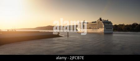 Kreuzfahrtschiff segelt auf einem ruhigen Fluss bei warmem Sonnenuntergang. Friedliches Wasser und eine malerische Küste schaffen eine elegante Reisetmosphäre. Stockfoto