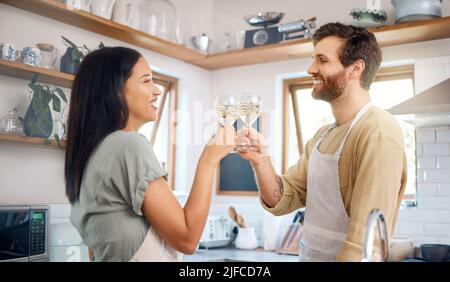 Junge interrassische Paare, die sich mit Weingläsern anstoßen, während sie Schürzen tragen und in der Küche zu Hause zusammen kochen. Junges Paar genießt romantisch Stockfoto