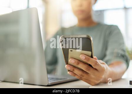 Nahaufnahme einer Frau mit gemischter Rasse, die ein Telefon in der Hand hält, während sie zu Hause an einem Laptop arbeitet. Eine hispanische Frau, die soziale Medien auf einem Mobiltelefon nutzt Stockfoto