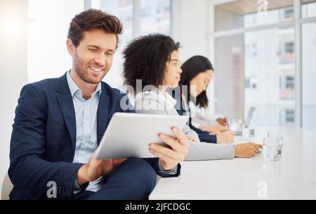 Der junge kaukasische Geschäftsmann, der während einer Präsentation in einem Sitzungssaal mit seinen verschiedenen Kollegen im auf einem digitalen Tablet-Gerät surft Stockfoto