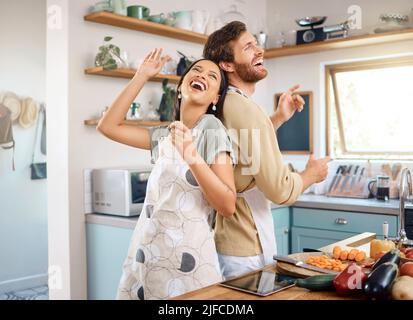 Fröhliches junges interrassisches Paar, das in der Küche spielerisch singt und tanzt, während es zusammen kocht. Glücklicher junger Mann und Frau in Schürzen Stockfoto