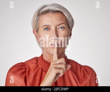 Eine reife kaukasische Frau, die mit einem Finger auf den Lippen stigulte, um ruhig zu sein und ein Geheimnis vor einem grauen Copyspace-Hintergrund zu bewahren. Alternde Frau, die huschend Stockfoto