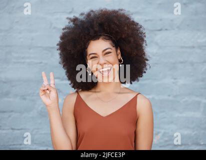 Eine junge Geschäftsfrau mit gemischter Rasse, deren Afro draußen gegen eine graue Wand steht und mit der Hand das Friedenszeichen gestikelt. Glücklich und zuversichtlich Stockfoto