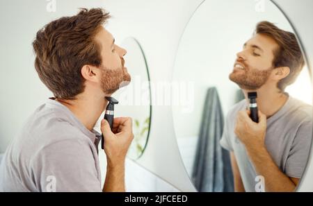 Ein hübscher Mann, der sich zu Hause im Badezimmer die Gesichtshaare rasiert. Kaukasischer Mann, der einen Rasierer benutzt, während er in einem Spiegel in seiner Wohnung aussiehen Stockfoto