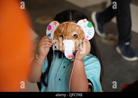 Ein kleines Mädchen, das eine farbige Tierpapiermaske vor ihrem Gesicht trägt, ist im Hintergrund des Klassenzimmers mit einem orangefarbenen Ballon isoliert Stockfoto