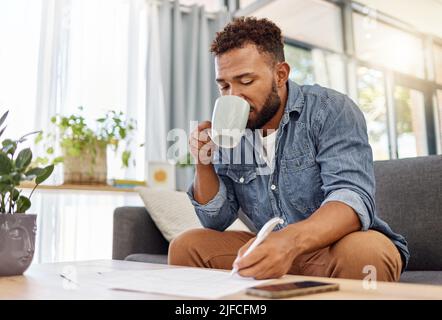 Junger Mann mit gemischter Rasse, der am Ausfüllen von Formularen arbeitet, während er zu Hause Kaffee trinkt. Eine hispanische Person, die eine Tasse Tee trinkt, während sie alleine in der Planung ist Stockfoto