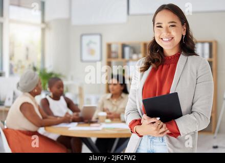 Lächelnde asiatische Geschäftsfrau mit einem digitalen Tablet, während Kollegen im Büro hinter ihr sitzen. Ehrgeiziges und glückliches professionelles Standing Stockfoto