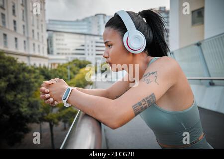 Junge seriöse Mixed Race hispanic-Sportlerin, die nachdenklich in Kopfhörern aussieht und Musik hört, während sie draußen auf einer Brücke steht Stockfoto