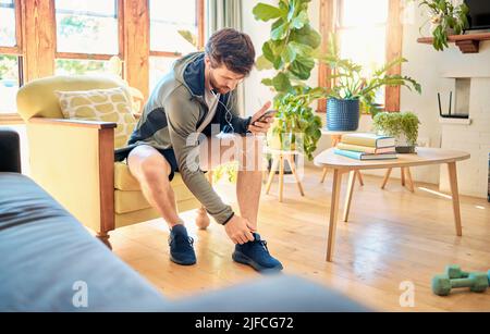 Ein junger Kaukasier, der sich zum Training anzieht, während er zu Hause mit Kopfhörern auf einem Mobiltelefon Musik hört. Kerl, der auf einem sitzt Stockfoto