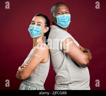 Covid geimpfte einen afroamerikanischen Mann und eine Frau mit gemischter Rasse, die Rücken an Rücken stand. Zwei Personen tragen eine chirurgische Gesichtsmaske, die gegen Rot isoliert ist Stockfoto