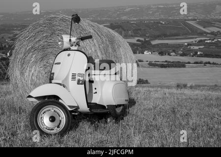 Bild eines alten Piaggio vespa Motorrads, das auf einem Feld mit Strohballen und einer atemberaubenden Aussicht im Hintergrund geparkt ist. Schwarzweißfotos. Stockfoto