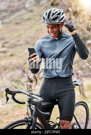 Afroamerikanischer Radfahrer trägt einen Helm, während er ein Handy benutzt und einen Sieg feiert. Männlich Freude beim Radfahren auf einem Fahrrad und beim Trainieren ausdrücken Stockfoto