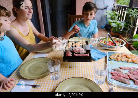 Familie beim Mittagessen auf der Terrasse ihres Hauses kocht die Frau das Fleisch auf einem heißen Stein. Stockfoto