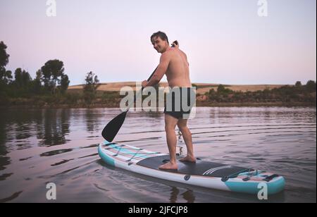 Junger aktiver Kaukasischer lächelt und nutzt sein Stand-Up-Paddle-Board auf einem See in der Natur. Junger Mann genießt einen Sommertag in der Morgendämmerung auf einem Paddel Stockfoto