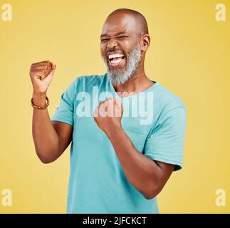 Ein reifer afrikanischer Mann, der beim Feiern ekstatisch aussieht, indem er mit den Händen eine Faustpumpenbewegung macht und gegen ein gelbes Studio jubelt Stockfoto