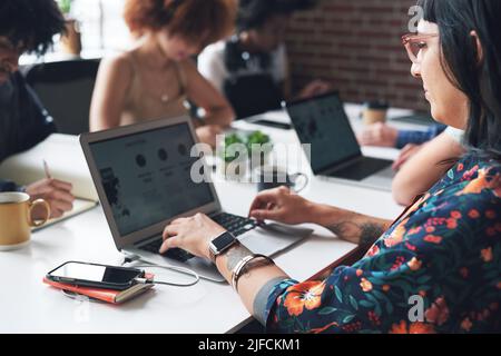 Eine junge Geschäftsfrau, die in ihrem Büro mit ihren Kollegen an ihrem Laptop arbeitet. Eine junge Geschäftsfrau trägt in einem Büro eine Brille, die auf ihrem Laptop tippt Stockfoto