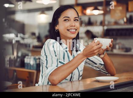 Fröhliche junge lächelnde Mischling Frau im lässigen Outfit, die heißes Getränk trinkt, während sie allein an einem Tisch im gemütlichen Café sitzt. Schöne weibliche aussehende Stockfoto