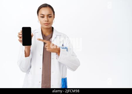 Besorgter Arzt zeigt mit dem Finger auf den Bildschirm des Mobiltelefons, runzelt die Stirn und sieht ernst aus, steht auf weißem Hintergrund. Online medizinische Klinik App Stockfoto