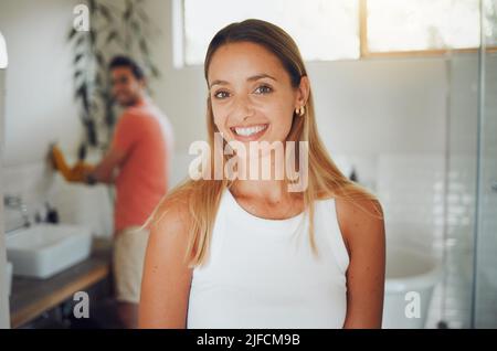 Das junge kaukasische Paar teilt sich ein Badezimmer mit Fokus auf eine fröhliche junge Frau, die lächelt, während sie auf die Kamera im Vordergrund schaut, während sie darauf wartet, zu kommen Stockfoto