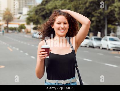 Porträt einer glücklichen, schönen jungen Frau mit gemischter Rasse, die die Stadt erkundet, während sie einen Kaffee zum Mitnehmen hält und ihr lockiges, brüniertes Haar berührt. Hispanisch Stockfoto