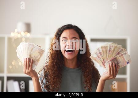 Schöne junge afroamerikanische Frau, die Stapel Papiergeld hält, während sie zu Hause in ihrem Wohnzimmer steht. Fröhliche Mischrennerin zeigt sich Stockfoto