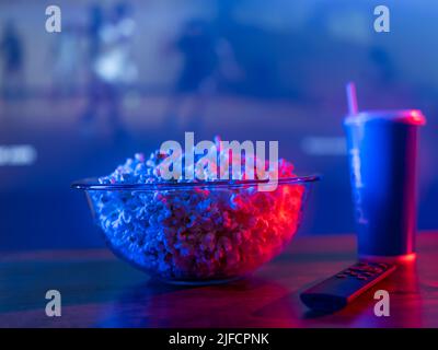 Auf einem Holztisch ein kohlensäurehaltiges Getränk in einem Plastikglas, eine Schüssel Popcorn vor dem Hintergrund eines großen Fernsehbildschirms. Sehen Sie Ihre Lieblingsfilme, Stockfoto