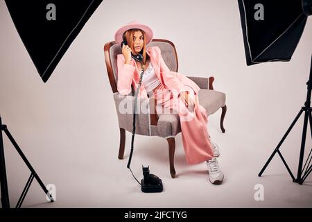 Stilvolle junge Mischling weibliche posiert in trendigen rosa Kleidung auf einem Stuhl sitzen und einen Anruf mit einem Retro-Telefon für ein Studio-Fotoshooting Stockfoto