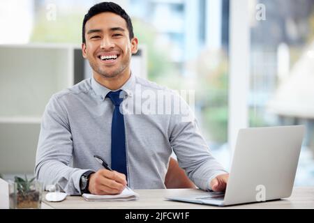 Porträt eines fröhlichen Geschäftsmannes mit gemischter Rasse, der in einem Tagebuch schreibt, während er allein im Büro an einem Laptop arbeitet. Ein Inhalt hispanic männlich Stockfoto