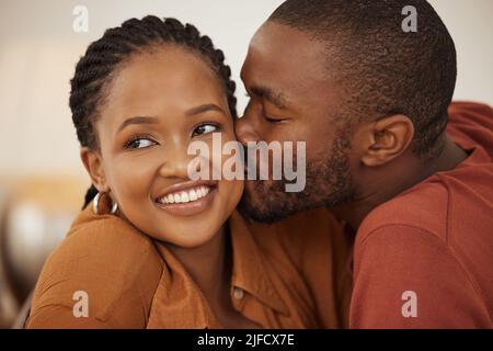 Liebevoller junger afroamerikanischer Mann, der seine Frau auf die Wange küsst, während sie lächelt und wegschaut. Glücklicher junger Mann und Frau teilen romantische intime Stockfoto