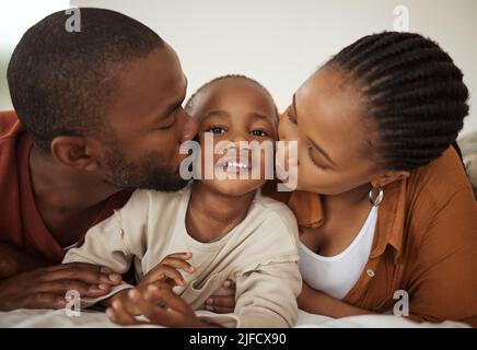 Porträt eines entzückenden kleinen afroamerikanischen Jungen, der zu Hause auf einem Bett zwischen Mutter und Vater liegt. Liebevolle Eltern küssen ihren kleinen Sohn Stockfoto