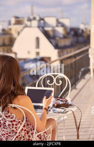 Folgen Sie mir für erstaunliche Pariser Sehenswürdigkeiten zu sehen. Aufnahme einer Frau mit Smartphone und Laptop beim Frühstück auf dem Balkon einer Wohnung Stockfoto