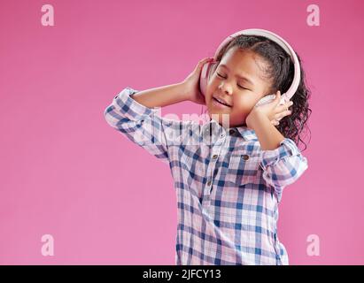 Ein hübsches kleines gemischtes Rennmädchen mit lockigem Haar, das Musik hört, während es Kopfhörer vor einem rosa Copyspace-Hintergrund in einem Studio trägt. Afrikanisch Stockfoto