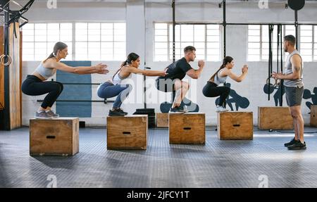 Eine vielfältige Gruppe aktiver junger Menschen, die gemeinsam mit einem Trainer in einem Fitnessstudio Boxjump-Übungen machen. Fokussierte Sportler landen dabei in der Hocke Stockfoto