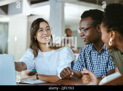 Drei junge, glückliche Geschäftsleute, die bei einer Besprechung an einem Tisch sitzen und bei der Arbeit an einem Laptop arbeiten. Business Professionals sprechen und Stockfoto