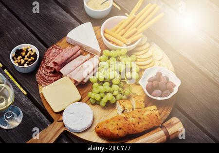 Blick über eine Vielzahl von Snacks auf einem Tapas-Holzbrett draußen. Käse, Brot, frische Trauben und Aufschnitt zum Mittagessen auf einem Weinberg. Essen Stockfoto