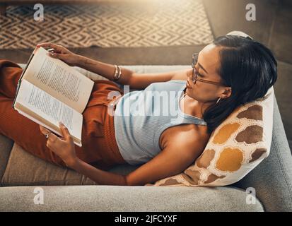 Oberhalb Ansicht einer Frau mit gemischter Rasse, die in einem Wohnzimmer zu Hause ein Buch liest. Wunderschöne hispanische Gäste, die alleine auf dem Loungesofa sitzen und Roman genießen. Entspannt Stockfoto
