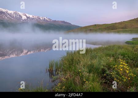 Erstaunlich zarte Morgendämmerung über den Bergen mit Schnee und Wald bedeckt, Nebel über dem See und schöne Reflexionen im Wasser gegen den blauen Himmel a Stockfoto