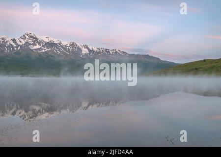 Erstaunlich zarte Morgendämmerung über den Bergen mit Schnee und Wald bedeckt, Nebel über dem See und schöne Reflexionen im Wasser gegen den blauen Himmel a Stockfoto