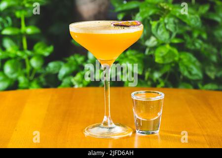 Eine Nahaufnahme eines Cocktails in einem Glas, das mit Passionsfrucht auf einem Tisch gegen grüne Pflanzen dekoriert ist Stockfoto